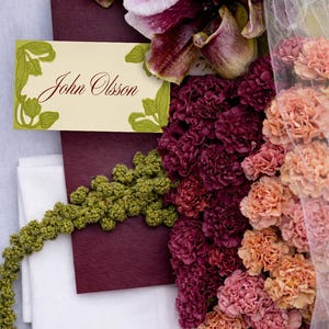 May include: A floral arrangement featuring orchids, carnations, and greenery. A cream-colored name card with the name "John Olsson" is visible. The flowers are in shades of burgundy, peach, and cream.