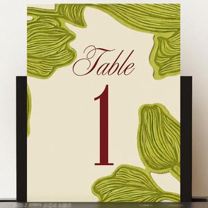 May include: A table number card with a cream background, featuring the word "Table" in a burgundy script font and the number "1" in a bold burgundy font. The card is decorated with green leaf-like designs.