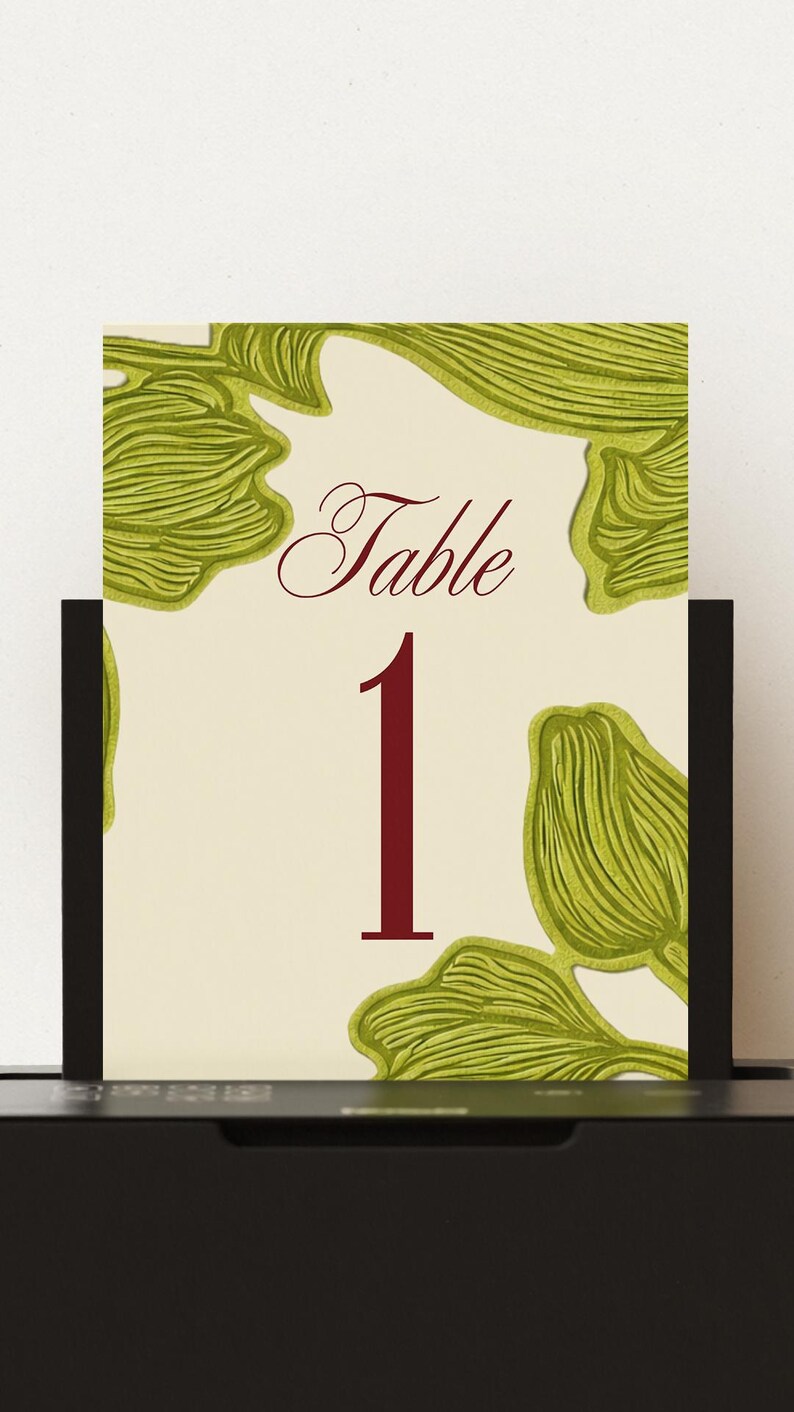 May include: A table number card with a cream background, featuring the word "Table" in a burgundy script font and the number "1" in a bold burgundy font. The card is decorated with green leaf-like designs.