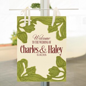 May include: A rectangular wedding welcome sign with a cream background and green floral border. The sign reads "Welcome TO THE WEDDING OF Charles & Haley 07.08.2026" in burgundy script. White ribbons are attached at the top for hanging.