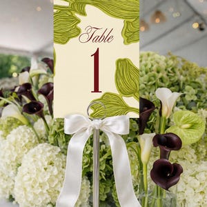 May include: A table number card with a cream background and green leaf design. The word "Table" and the number "1" are in burgundy. The card is held by a silver stand with a white satin bow. The background features white and green flowers.