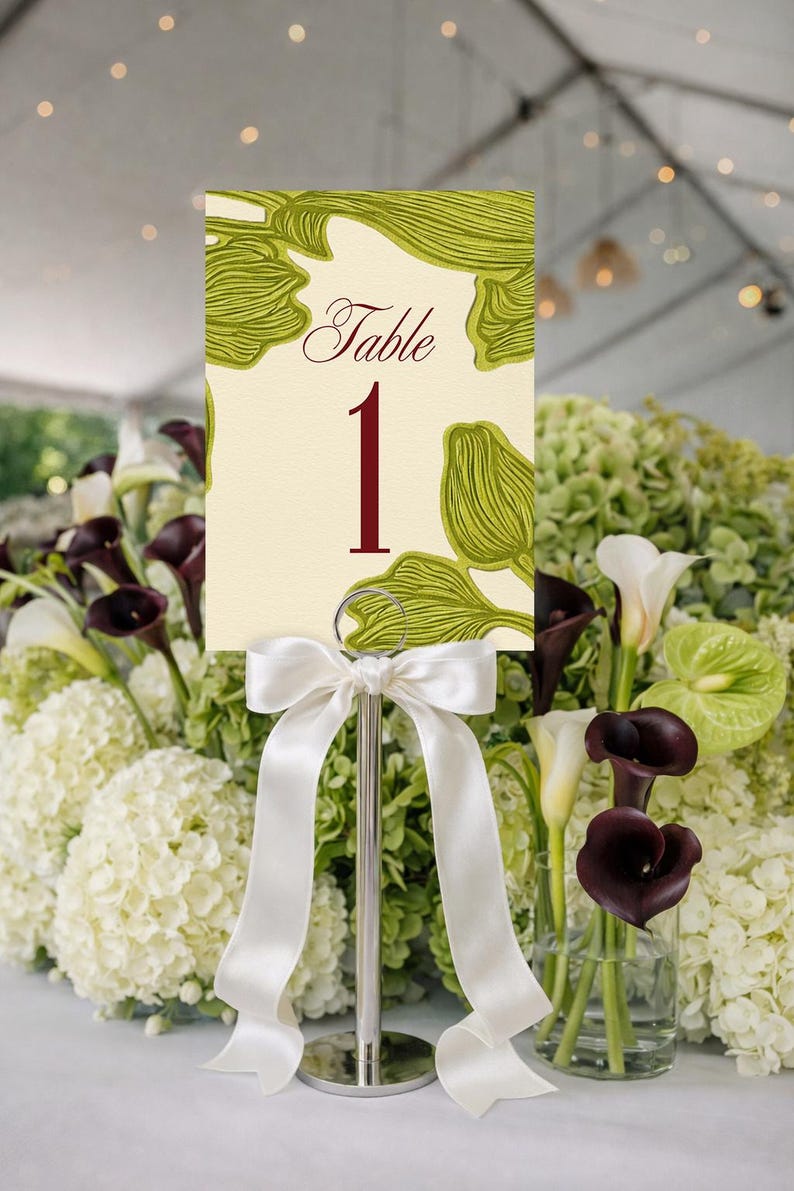 May include: A table number card with a cream background and green leaf design. The word "Table" and the number "1" are in burgundy. The card is held by a silver stand with a white satin bow. The background features white and green flowers.