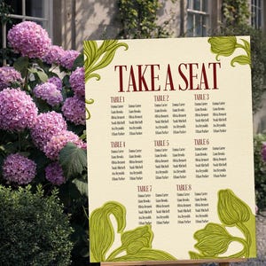 May include: A cream-colored wedding seating chart with the words "TAKE A SEAT" in burgundy. The chart lists names under table numbers 1-8, with green floral accents. It is displayed on a wooden easel outdoors.