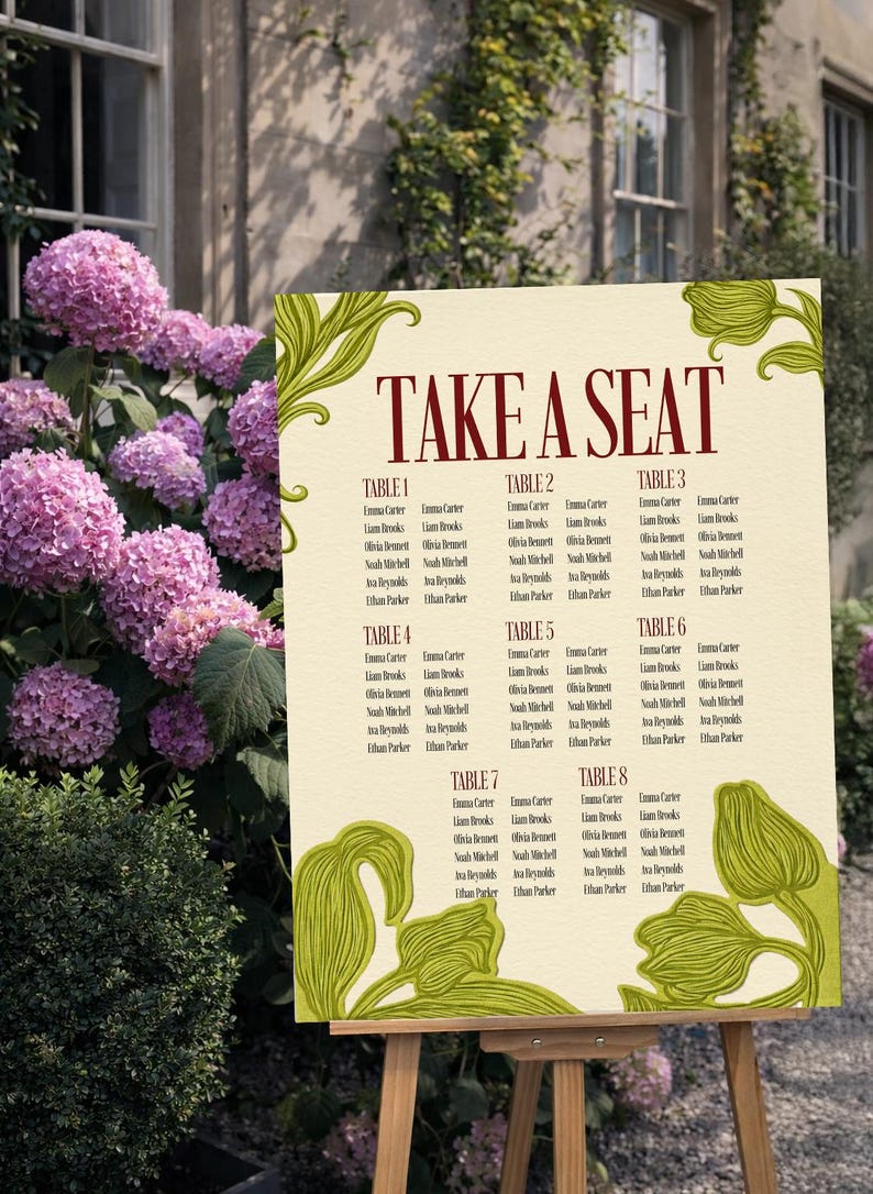 May include: A cream-colored wedding seating chart with the words "TAKE A SEAT" in burgundy. The chart lists names under table numbers 1-8, with green floral accents. It is displayed on a wooden easel outdoors.