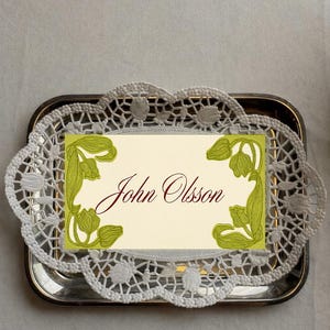 May include: A silver tray holds a cream-colored card with the name "John Olsson" in burgundy script. The card is framed with green floral illustrations and rests on a white lace doily.