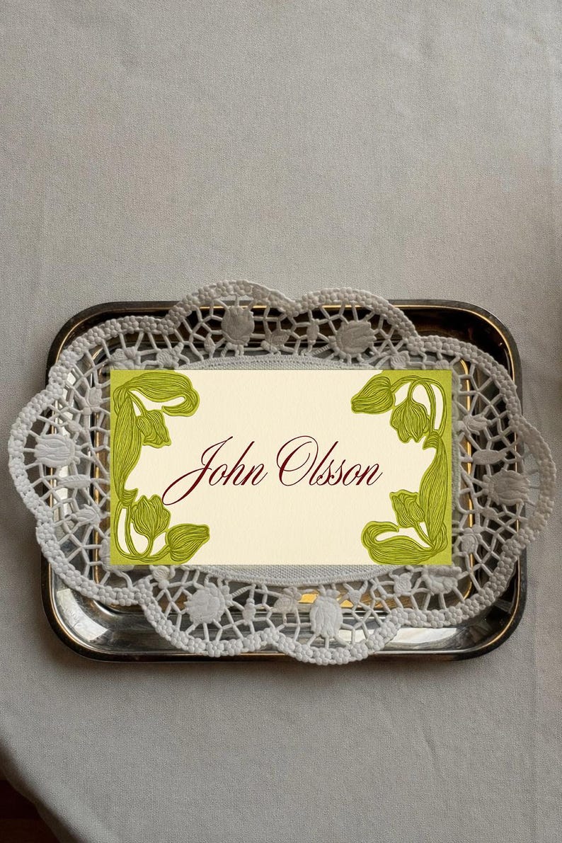May include: A silver tray holds a cream-colored card with the name "John Olsson" in burgundy script. The card is framed with green floral illustrations and rests on a white lace doily.