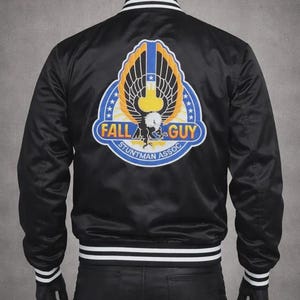 May include: Black satin bomber jacket with white and black striped trim. The back features a colorful patch with the words "FALL GUY STUNTMAN ASSOC" and a stylized eagle design. The jacket has a classic bomber style with a ribbed collar and cuffs.