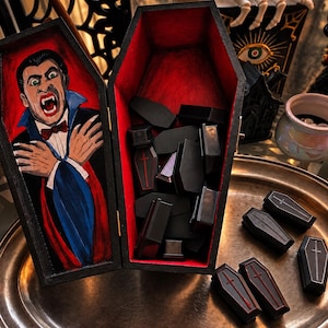 May include: A large black wooden coffin box with a red interior, featuring a hand-painted vampire on the lid. The open box contains several small black coffin-shaped containers with red cross details, and more are arranged on a metal tray nearby.