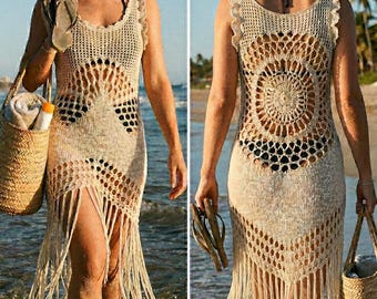 May include: A beige, sleeveless crochet dress with fringe detail. The dress features a round, openwork design on the back and a fringed hemline. The person is carrying a straw tote bag and sandals, walking on a beach.