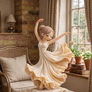 Op de afbeelding: Een gehaakte ballerinafiguur in een vloeiende crèmekleurige jurk, met sierlijk uitgestrekte armen. De pop heeft een knotje en staat op een vierkante basis. De achtergrond omvat een raam, een stoel en een lamp.