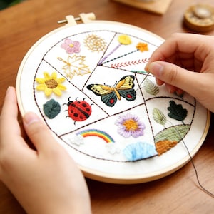 Può includere: Un telaio da ricamo rotondo con un disegno floreale e di insetti. Il disegno comprende un girasole giallo, una coccinella rossa, una farfalla, un arcobaleno e altri elementi colorati. Il ricamo è in lavorazione con ago e filo.