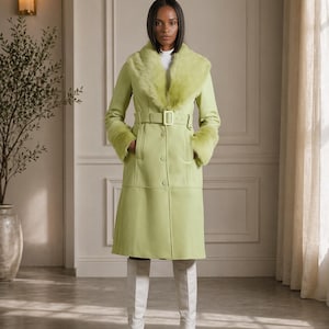 Peut inclure: Un manteau vert clair avec un col et des poignets en fausse fourrure. Le manteau est ceinturé à la taille et porté avec des bottes blanches montantes. Le manteau a un devant boutonné et une jupe évasée. Un col roulé blanc est porté.
