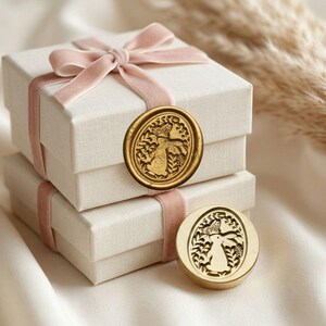 May include: Two stacked cream gift boxes tied with pink velvet ribbon and bow. A gold wax seal with a rabbit and butterfly design is on the top box. Another seal is below. The background is a soft, neutral colour.