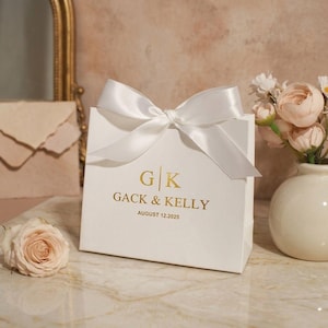 May include: A white gift bag with a satin bow. The bag features gold lettering: "G | K GACK & KELLY AUGUST 12.2025". A vase of flowers and a rose are in the background. The bag rests on a marble surface.