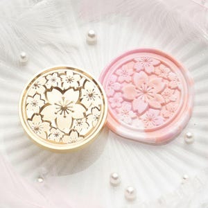 May include: Two circular wax seal stamps featuring cherry blossom patterns. One is gold-coloured, the other is pink and white. The stamps rest on a white surface with pearls and feathers.