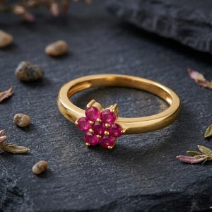 May include: A gold ring with a flower-shaped cluster of bright red gemstones. The ring is displayed on a dark surface, with small pebbles and leaves scattered around, emphasizing the ring's design and colour.