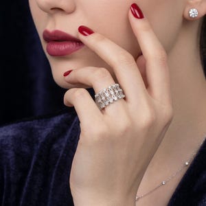 May include: A close-up of a hand wearing a silver ring with a repeating openwork design, accented with small, sparkling stones. The ring is on the ring finger. The person is also wearing a diamond stud earring and a delicate necklace.