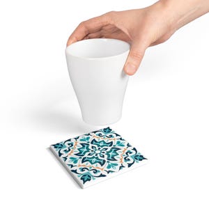 May include: A white ceramic cup held above a square coaster. The coaster features a teal, navy, and orange floral pattern on a white background. The cup is smooth and cylindrical.