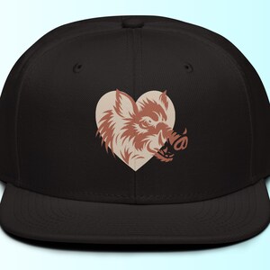 Puede incluir: Gorra de béisbol negra con un diseño en forma de corazón. El diseño presenta una ilustración detallada de un jabalí en tonos marrones y beige. La gorra tiene una visera plana y un ajuste estructurado.