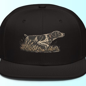 Puede incluir: Gorra negra de ala plana con una ilustración de un perro en un campo de hierba alta. El perro está representado en tonos marrones y negros, con un collar marrón. La gorra es de estilo clásico, adecuada para actividades al aire libre.
