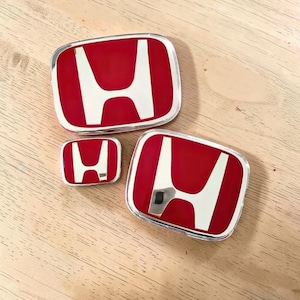 Könnte beinhalten: Drei Honda-Fahrzeugembleme mit einem leuchtend roten Hintergrund und einem metallisch silbernen H-Logo. Die Embleme sind rechteckig mit abgerundeten Ecken und einem polierten, chromartigen Rand und zeigen deutlich die Identität der Automarke.