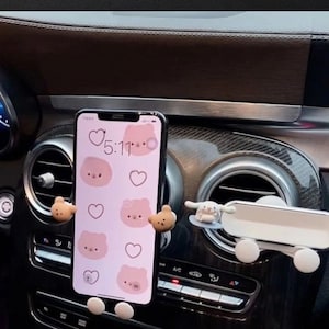 May include: A car phone holder with a smartphone displaying a pink bear-themed wallpaper. The holder features cute bear-shaped grips and a white, adjustable mount. The dashboard has a dark wood and silver trim.
