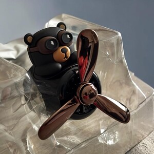 Airplane car air freshener with rotating propeller and pilot bear design, designed as a vent clip diffuser for car interiors. This decorative car accessory provides light fragrance while adding a fun aviation-inspired touch. Made from durable ABS material, it easily attaches to air vents and spins with airflow. Perfect for enhancing car decor and keeping your vehicle fresh during daily drives or road trips.