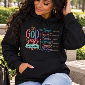 Puede incluir: Una sudadera negra con capucha que presenta un estampado gráfico colorido con el texto "GOD says you are Unique, Special, Lovely, Precious, Chosen, Forgiven" y varias referencias bíblicas como Salmos 139:13 y Juan 15:16.