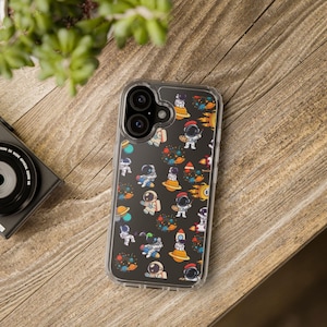 Puede incluir: Funda transparente para teléfono con un patrón repetido de astronautas de dibujos animados, planetas y cohetes. Los astronautas están en varias poses y usan cascos y trajes de diferentes colores. La funda está sobre una superficie de madera.