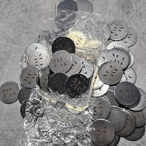 May include: A collection of circular dark metal tokens featuring the engraved text IL BORRO, PK, and 5.23.26, partially contained within a clear plastic box wrapped in bubble wrap, with several loose tokens scattered around the container.