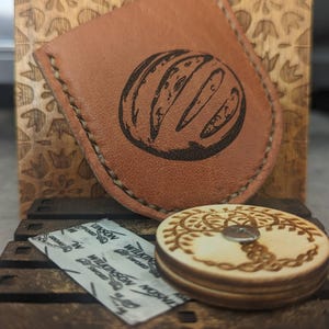 Puede incluir: Un estuche de cuero marrón con un diseño gráfico negro, un posavasos de madera con un diseño decorativo y un paquete pequeño. El estuche y el posavasos están sobre una superficie de madera, con un fondo de madera tallada.