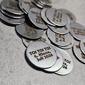 Op de afbeelding: Een verzameling ronde, zilverkleurige metalen penningen die in een hoopje verspreid liggen. Elke penning is voorzien van gegraveerde zwarte tekst met de tekst TOI TOI TOI, 6. Klasse, Juli 2026, terwijl andere een kleine afbeelding van een kasteel en de naam Robin Hood tonen.