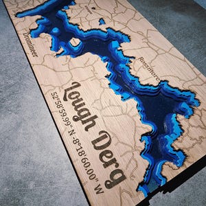 May include: A wooden map of Lough Derg, showing layered blue and dark blue water depths. The map includes the text "Lough Derg" and coordinates 52°58'59.99" N -8°18'60.00" W. The map is on a light brown wooden surface.
