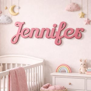 May include: A pink wooden sign that spells out the name "Jennifer" in a cursive font, mounted on a light pink wall. The room is decorated with a crib, a dresser, and hanging cloud, moon, and star decorations.
