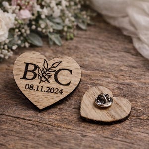 Op de afbeelding: Twee houten hartvormige spelden. Eén speld heeft de initialen "B&C" met een bloemmotief en de datum "08.11.2024" gegraveerd. De andere speld heeft een metalen sluiting aan de achterkant. De spelden liggen op een houten oppervlak.