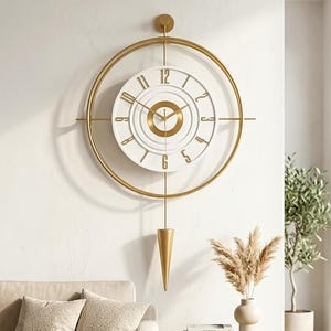 Könnte beinhalten: Eine Wanduhr mit einem großen goldenen kreisförmigen Rahmen, der ein weißes Zifferblatt mit goldenen Ziffern umschließt. Die Uhr hat einen zentralen goldenen Ring, goldene Zeiger und ein goldenes konisches Pendel.