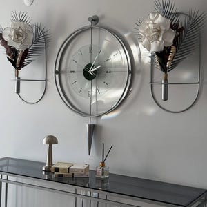 May include: A modern wall decor arrangement featuring a large, round, silver-framed clock with a glass face. Two matching wall sconces hold white flower arrangements. A console table below displays a lamp, books, and a fragrance diffuser.