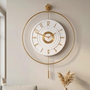 Puede incluir: Un reloj de pared circular con esfera blanca, números romanos dorados y un disco central texturizado en color oro. El reloj está enmarcado por un fino anillo de metal dorado con una varilla vertical y un peso de péndulo colgante.