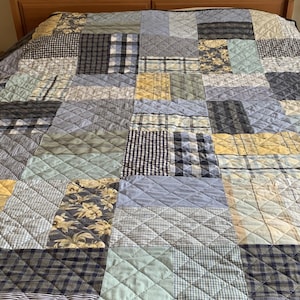 May include: A quilted bedspread featuring a patchwork design of rectangular fabric pieces in shades of blue, yellow, and white. The pattern includes various textures such as gingham, plaid, stripes, and floral prints, all stitched together with a diamond-shaped quilting pattern.