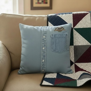 Peut inclure: Un coussin décoratif bleu clair avec un motif de chemise en jean. Le coussin présente un devant boutonné, une poche en jean et un emblème brodé. Il est placé sur un canapé beige avec une courtepointe à motifs.