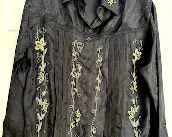May include: Black button-down shirt with floral embroidery in gold and silver. The shirt features a collared neckline, vertical pleats, and long sleeves. The floral embroidery is on the collar, front, and sleeves. The shirt is hanging on a hanger.
