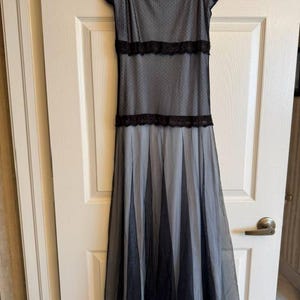 May include: A sleeveless midi dress featuring a light gray base layer with a black polka dot mesh overlay. The dress includes black lace trim along the neckline and two horizontal bands across the bodice, with a pleated skirt bottom.