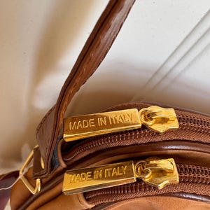 May include: Close-up of a brown leather crossbody bag with gold-toned zippers. The bag features the text "MADE IN ITALY" on the zipper pulls. The bag has a dual-compartment design and a brown leather strap.