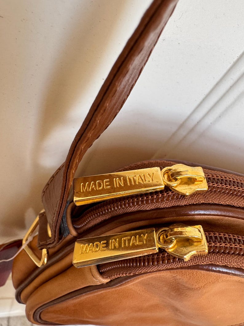 May include: Close-up of a brown leather crossbody bag with gold-toned zippers. The bag features the text "MADE IN ITALY" on the zipper pulls. The bag has a dual-compartment design and a brown leather strap.