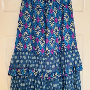 Puede incluir: Una falda larga y fluida con un estampado geom&eacute;trico azul, rosa y amarillo. La falda presenta una cintura de ganchillo y volantes en capas en la parte inferior. La falda est&aacute; colgada en una percha negra.
