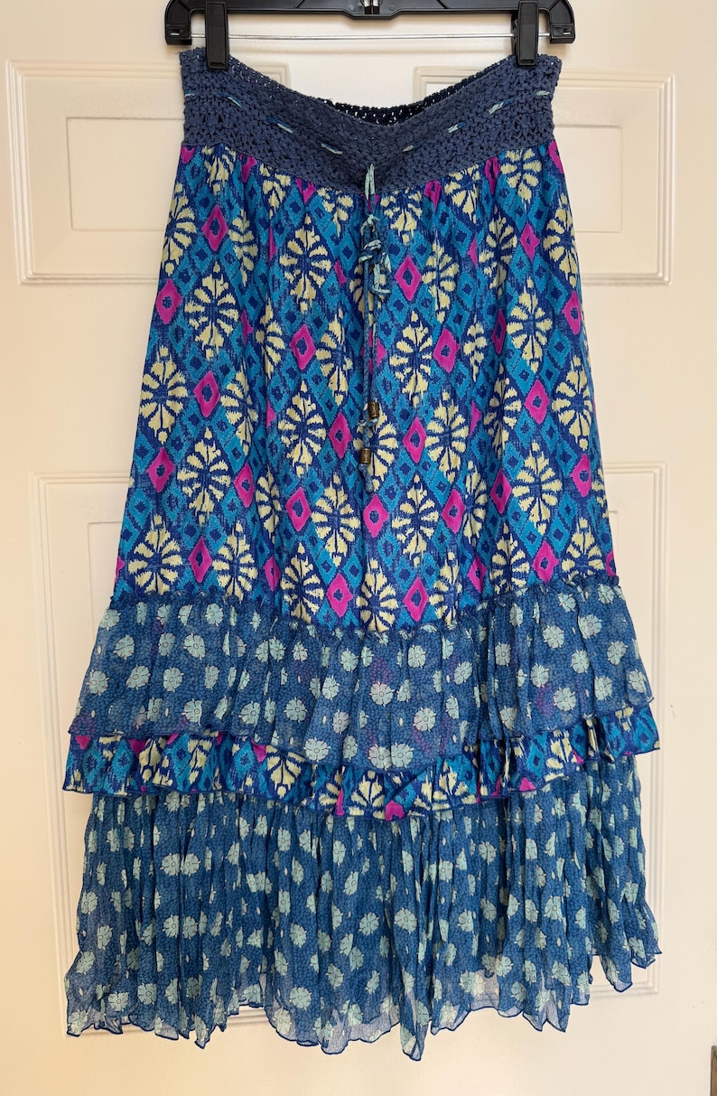 Puede incluir: Una falda larga y fluida con un estampado geom&eacute;trico azul, rosa y amarillo. La falda presenta una cintura de ganchillo y volantes en capas en la parte inferior. La falda est&aacute; colgada en una percha negra.