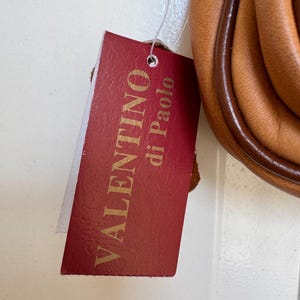 May include: A red tag with the words "VALENTINO di Paolo" in gold lettering is attached to a tan leather bag. The bag has a brown trim and a rounded shape. The tag is rectangular with a small hole at the top.
