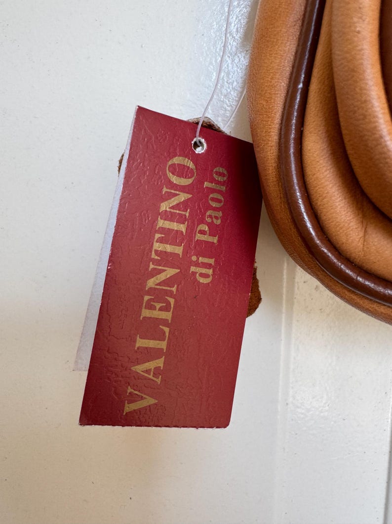 May include: A red tag with the words "VALENTINO di Paolo" in gold lettering is attached to a tan leather bag. The bag has a brown trim and a rounded shape. The tag is rectangular with a small hole at the top.