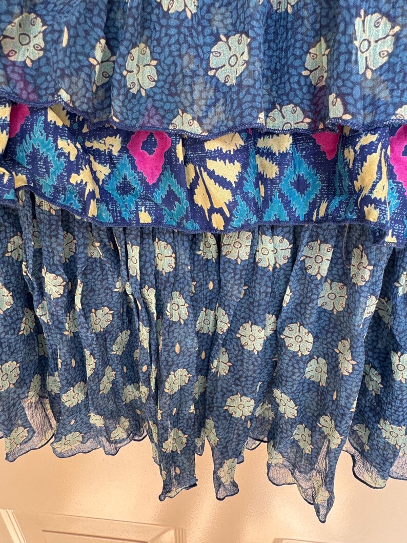 Puede incluir: Primer plano de una prenda fluida y escalonada en tonos azules. La tela presenta estampados florales y geom&eacute;tricos en azul claro, turquesa y magenta. Las capas crean un efecto de volantes, con una textura delicada y transparente.
