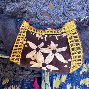 Puede incluir: Primer plano de un top de ganchillo azul con un parche estampado floral. El parche es de color p&uacute;rpura oscuro con flores blancas y ribete amarillo. La parte superior tiene un cord&oacute;n y est&aacute; hecha de varias telas azules.